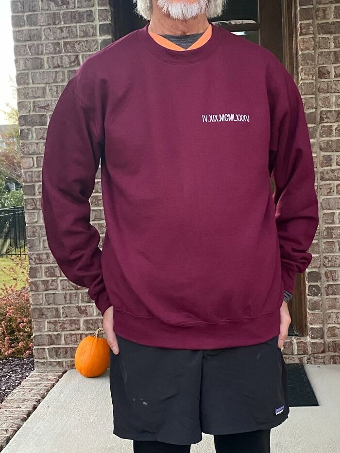 Man wearing a custom maroon sweatshirt with roman numerals, standing outside near a brick wall and a pumpkin for Christmas gifts.