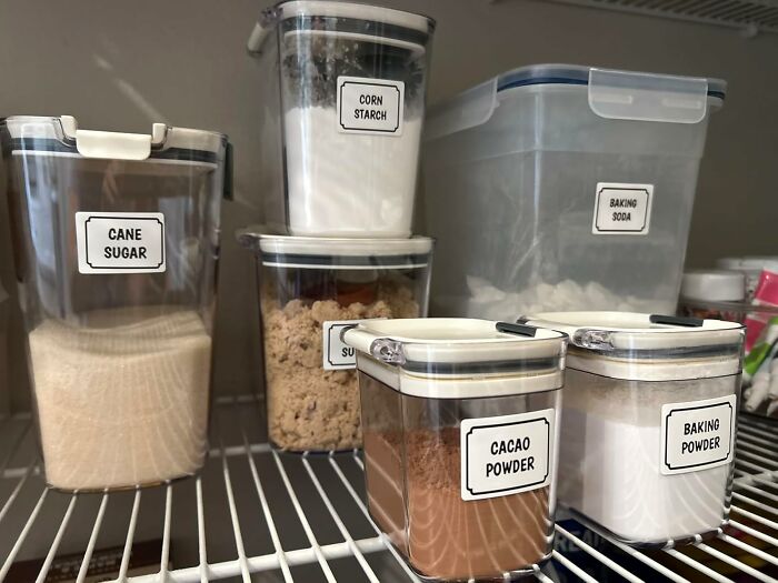 Clear labeled containers of baking ingredients including cacao powder, cane sugar, and baking soda for gift ideas.