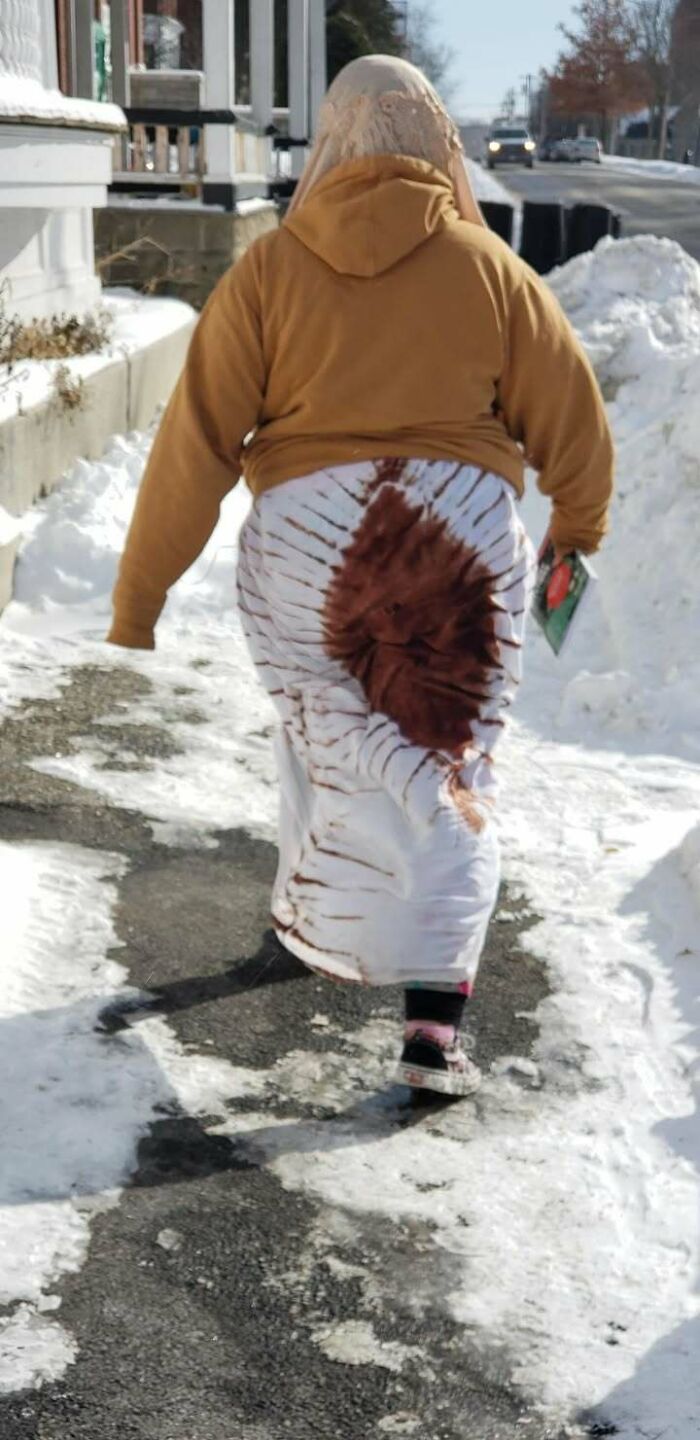 Person walking on snowy sidewalk wearing a brown hoodie and a skirt with a large random stain in a funny unexpected moment.