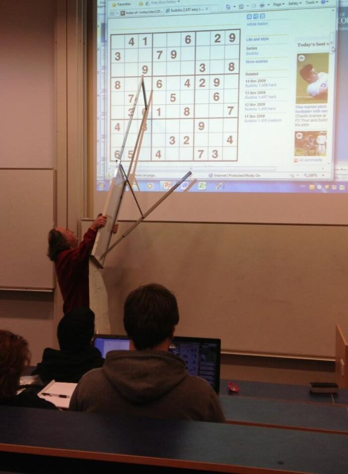 Professor humorously holding a broken desk while pointing at a projected Sudoku puzzle in a classroom setting.