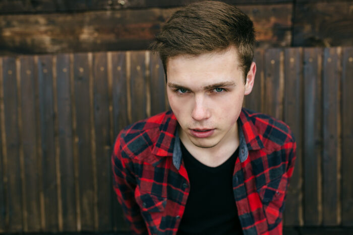 Young man in a red plaid shirt showing an expression of entitlement and frustration against a wooden background