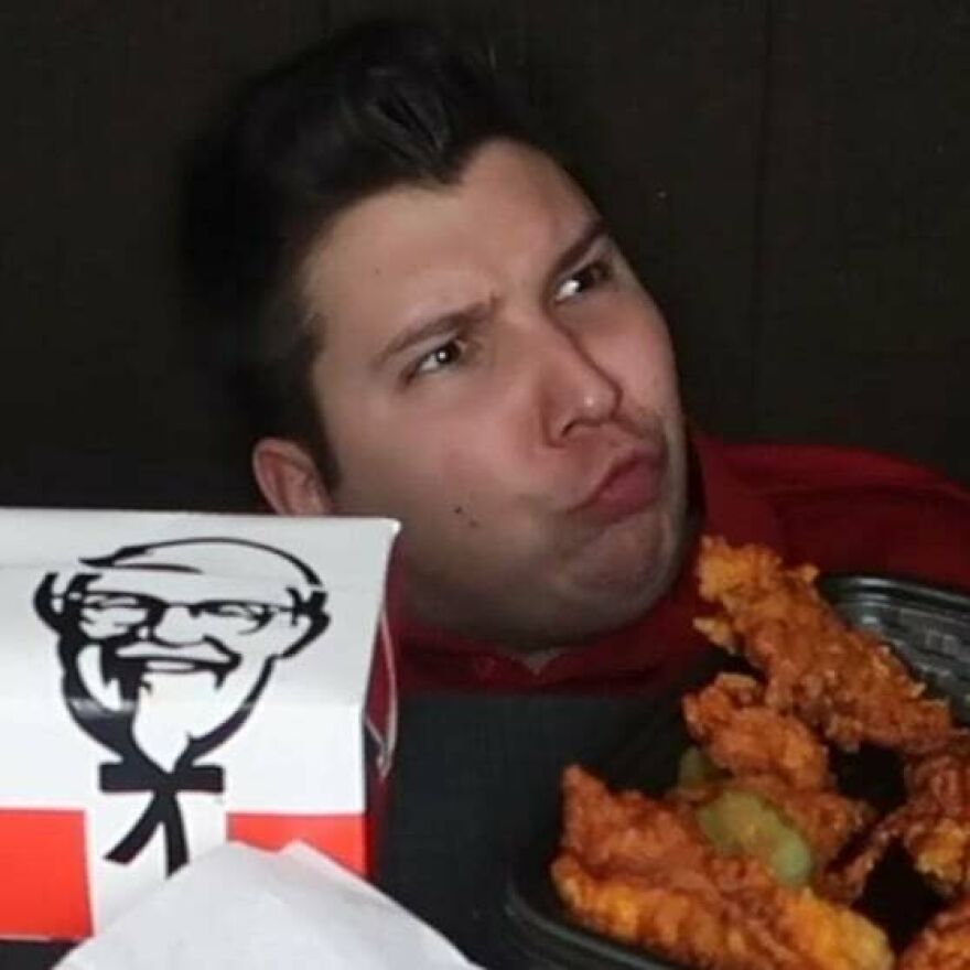 Nikocado Avocado posing with fried chicken and a KFC box, making a playful facial expression in a dark setting.
