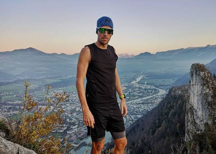 Man wearing sunglasses and athletic clothes standing on an Austrian mountain overlooking a valley and river below.
