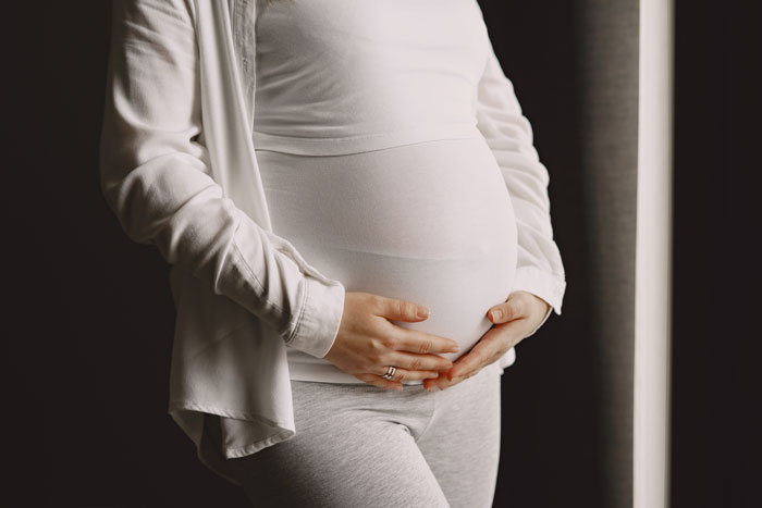 Pregnant woman in casual clothes gently holding belly near window with soft natural light, symbolizing upcoming kids.