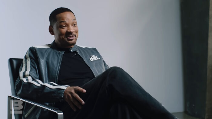 Man wearing a sporty jacket sitting in a chair, smiling during an interview about celebrities people think suffer.
