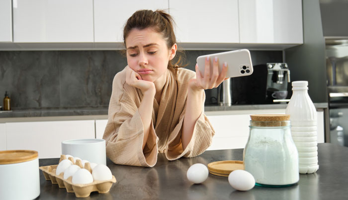 Woman at kitchen counter looking frustrated holding phone, dealing with picky eater and allergy concerns. Woman at kitchen counter looking frustrated holding phone, dealing with picky eater and allergy concerns.