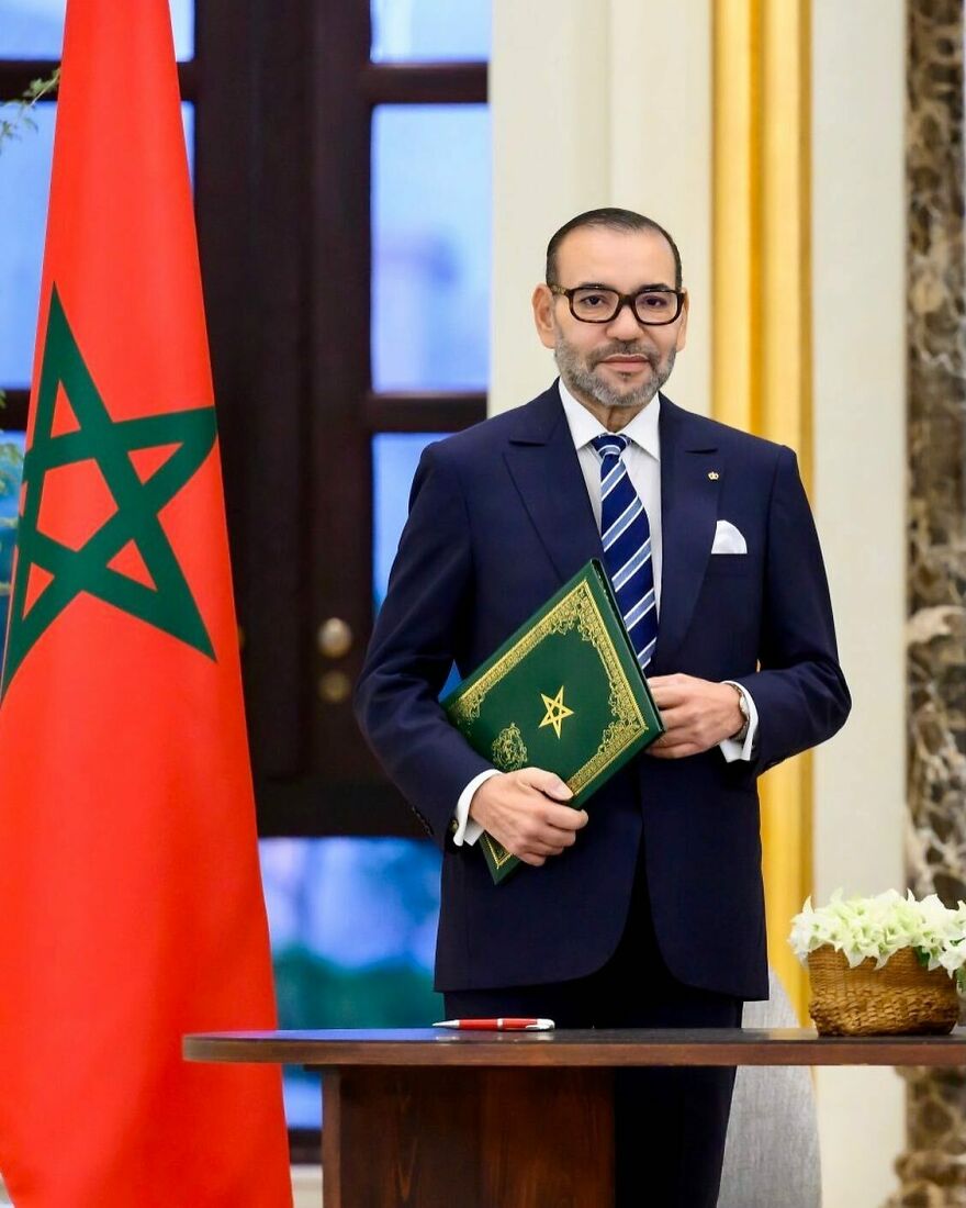 King of Morocco standing beside the national flag holding a green book, representing one of the oldest surviving monarchies.