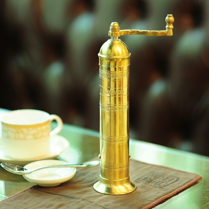 Brass pepper grinder on a table next to a cup and spoon, a unique gift for the person who has everything.