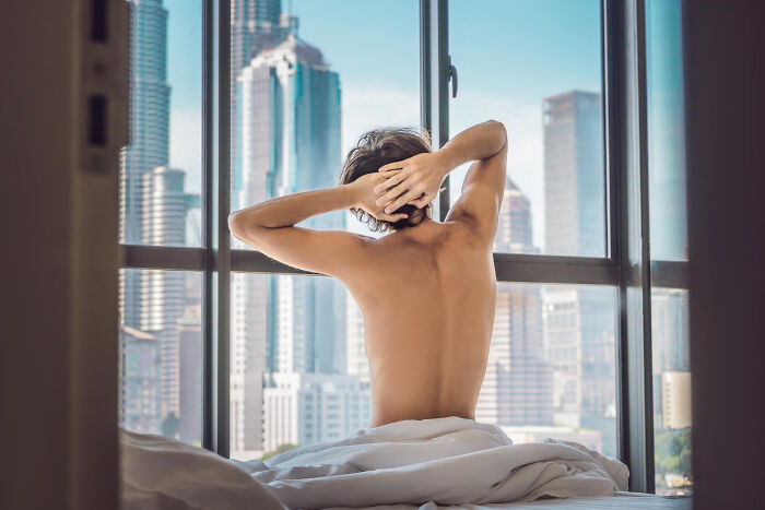 Person seen from behind through a window, stretching in bed with city skyline outside, bizarre neighbor window view.