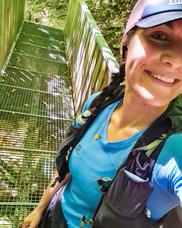 Smiling woman with a braided ponytail wearing hiking gear on a metal walkway in a forested Austrian mountain area.