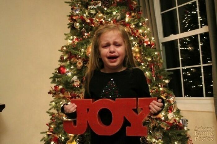 Young girl crying holding bright red JOY sign in front of decorated Christmas tree in awkward family Christmas photo.