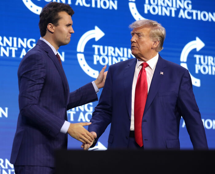 Charlie Kirk and Donald Trump shaking hands onstage at a Turning Point Action event with blue branded background. Charlie Kirk and Donald Trump shaking hands onstage at a Turning Point Action event with blue branded background.