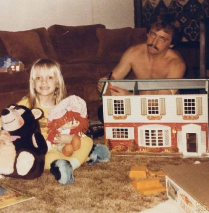 Awkward family Christmas photo of a shirtless man and smiling child with toys in a vintage living room setting.