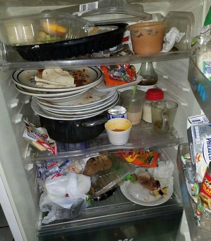 Messy dude fridge packed with dirty dishes, food containers, and snacks stacked on multiple shelves inside the refrigerator.