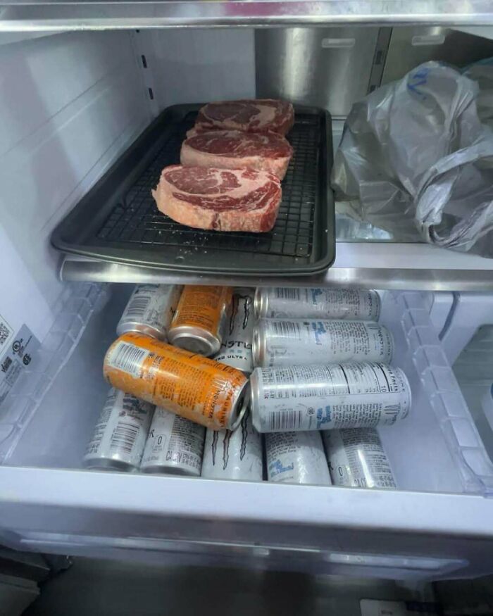 Refrigerator with raw steaks on a tray and multiple energy drink cans inside, showcasing a wild dude fridge setup.
