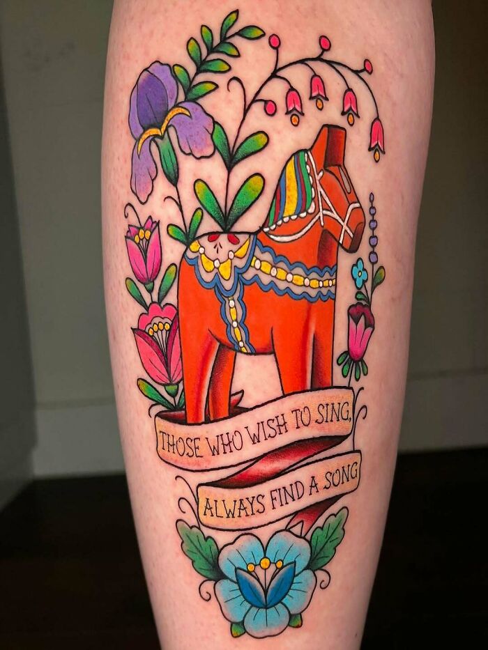Colorful wholesome tattoo of a traditional horse surrounded by flowers and a banner with a meaningful quote on the leg. Colorful wholesome tattoo of a traditional horse surrounded by flowers and a banner with a meaningful quote on the leg.