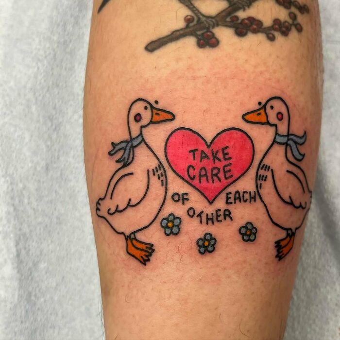 Wholesome tattoo of two ducks facing each other with a heart reading take care of each other and small flowers beneath.