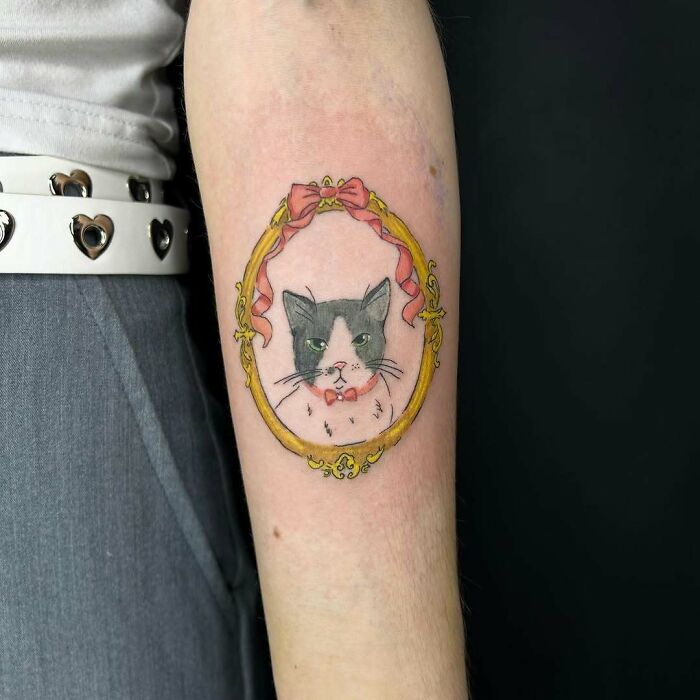 Tattoo of a gray and white cat wearing a bow tie inside a decorative gold frame with a pink ribbon on a forearm.