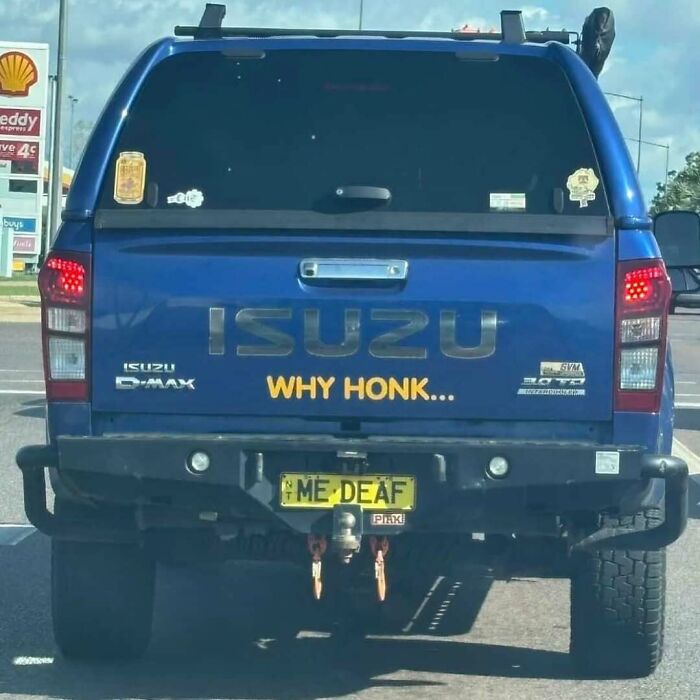 Blue Isuzu truck with a hilarious personalized license plate reading ME DEAF, parked near a gas station.