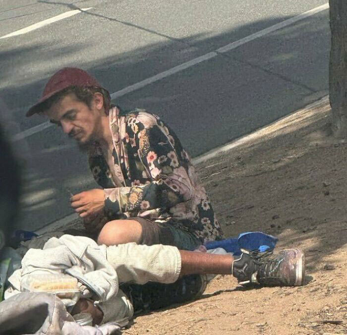 A man in a floral shirt and red cap sitting on the ground next to a person lying down, related to Tylor Chase hospitalization. A man in a floral shirt and red cap sitting on the ground next to a person lying down, related to Tylor Chase hospitalization.