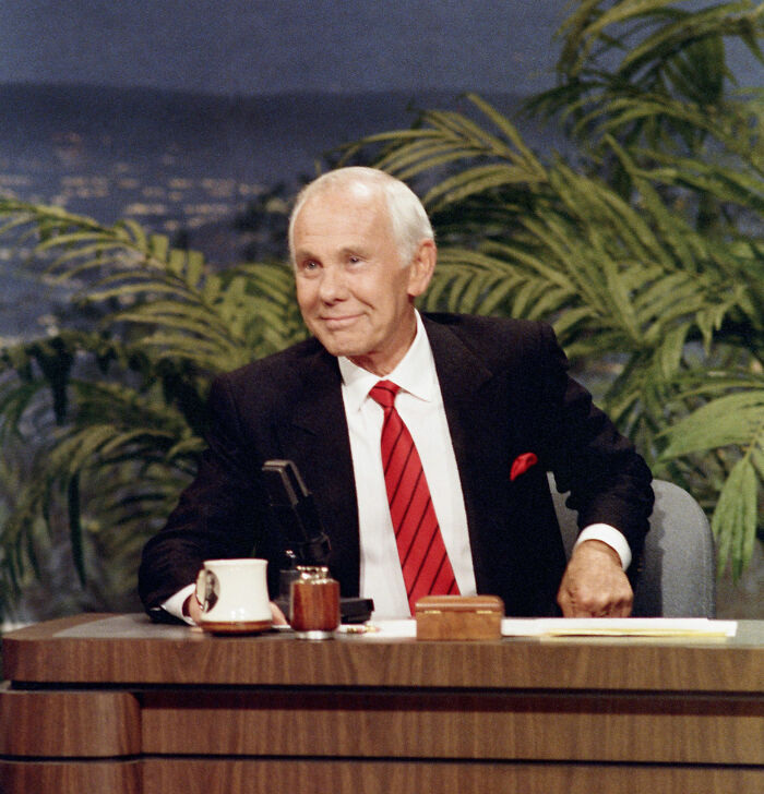 Johnny Carson hosting the Tonight Show, seated at the desk with a microphone and plants in the background. Johnny Carson hosting the Tonight Show, seated at the desk with a microphone and plants in the background.