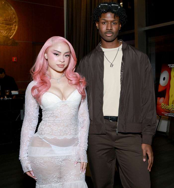 Ice Spice in a see-through lace dress with pink hair, standing next to a man in a brown jacket at an event. Ice Spice in a see-through lace dress with pink hair, standing next to a man in a brown jacket at an event.
