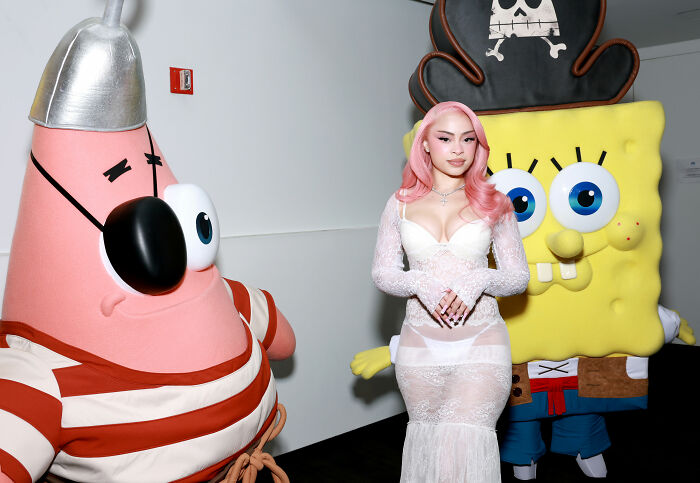 Ice Spice in a white see-through dress posing between Patrick Star and SpongeBob SquarePants characters at an event. Ice Spice in a white see-through dress posing between Patrick Star and SpongeBob SquarePants characters at an event.