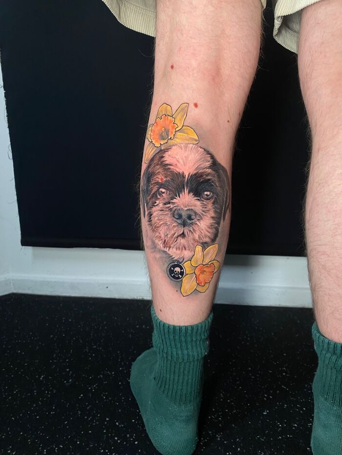 Realistic tattoo of a dog's face with yellow flowers on a person's lower leg showcasing pet tribute tattoos.