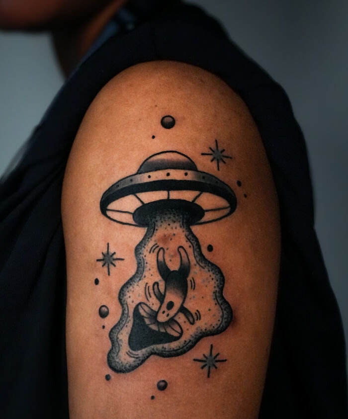 Black and gray psychedelic tattoo of a UFO abducting a creature surrounded by stars on a person's upper arm. Black and gray psychedelic tattoo of a UFO abducting a creature surrounded by stars on a person's upper arm.