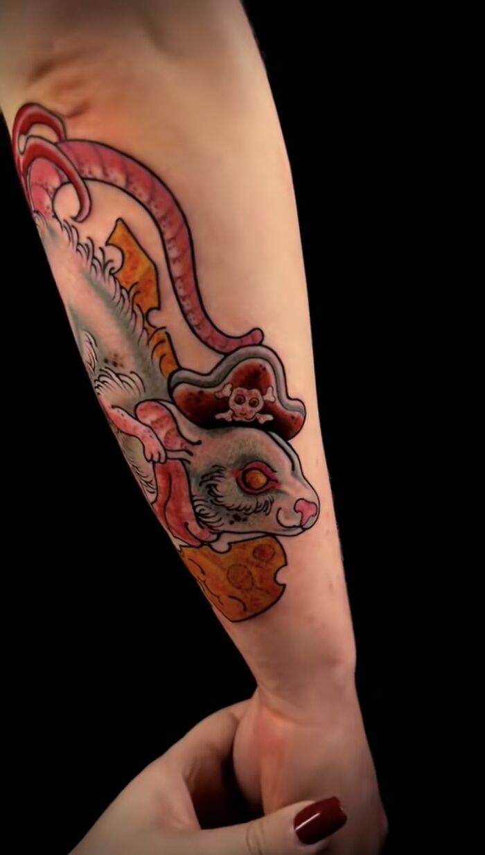 Colorful pet tribute tattoo of a rat wearing a pirate hat holding cheese on a forearm, showcasing pet memory art.
