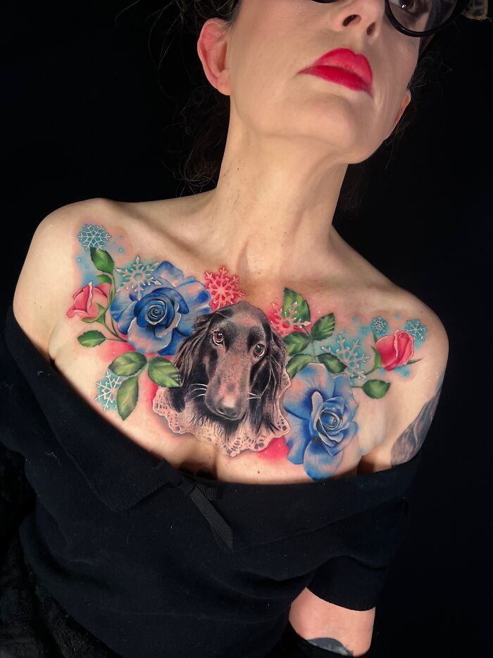 Woman showing colorful chest tattoo of a dog surrounded by blue roses, a tribute tattoo honoring pet memories.