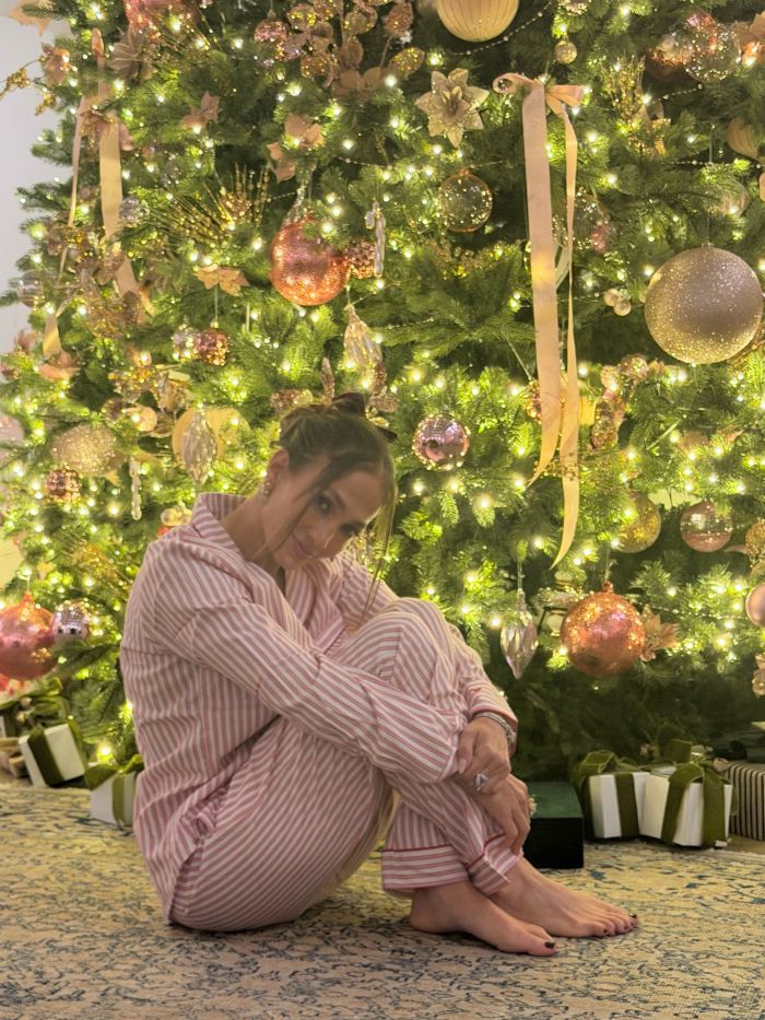 Jennifer Lopez sitting in striped pajamas by a decorated Christmas tree with lights and ornaments.