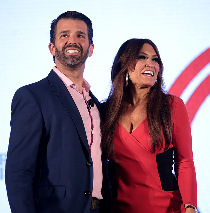 Donald Trump Jr. and Kimberly Guilfoyle smiling together on stage with bright background during event.