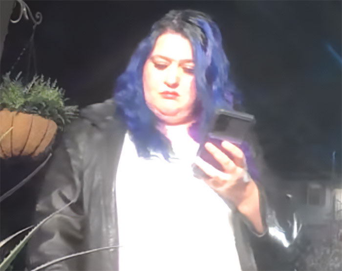 Person with blue hair and black jacket looking at a phone in a dark setting related to DoorDasher pepper-spraying incident.