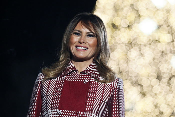 Melania Trump smiling in a red patterned coat with blurred warm lights in the background, linked to Christmas ornament outrage. Melania Trump smiling in a red patterned coat with blurred warm lights in the background, linked to Christmas ornament outrage.