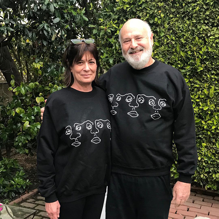 Couple wearing matching black sweatshirts with abstract faces, outdoors by green leafy bushes and brick path.