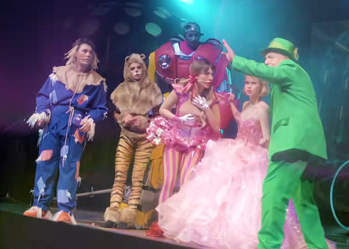 Cast members in colorful Wizard of Oz costumes performing on stage during a controversial theatrical production.