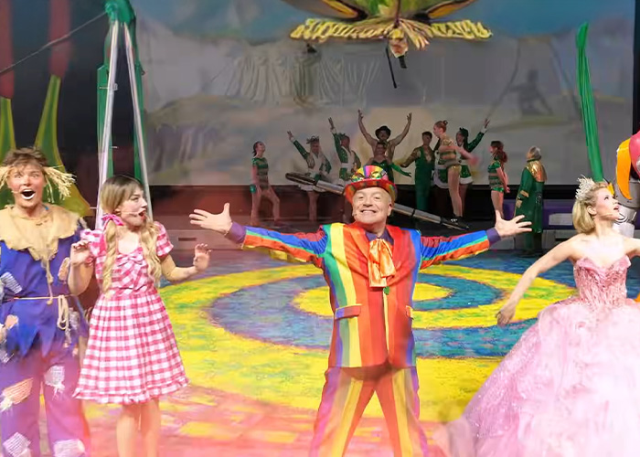 Actors in colorful costumes performing a Wizard of Oz scene on stage with vibrant lighting and a scenic backdrop. Actors in colorful costumes performing a Wizard of Oz scene on stage with vibrant lighting and a scenic backdrop.