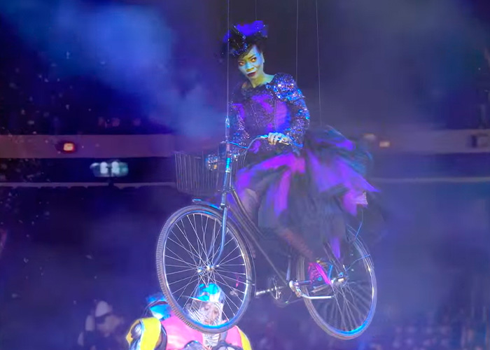 Performer in colorful costume on a suspended bicycle during Wizard of Oz performance amid audience reaction. Performer in colorful costume on a suspended bicycle during Wizard of Oz performance amid audience reaction.