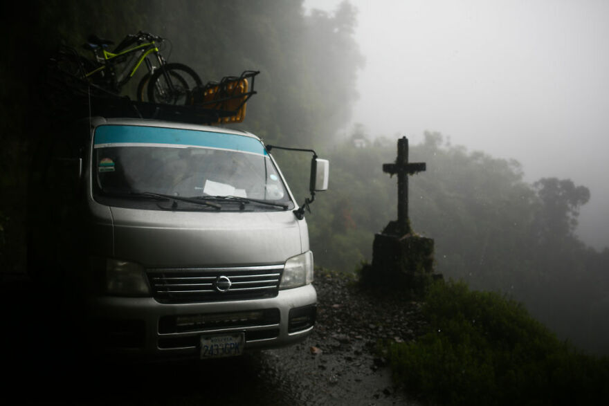 A van parked on a narrow foggy mountain road near a roadside memorial in a dangerous travel location.