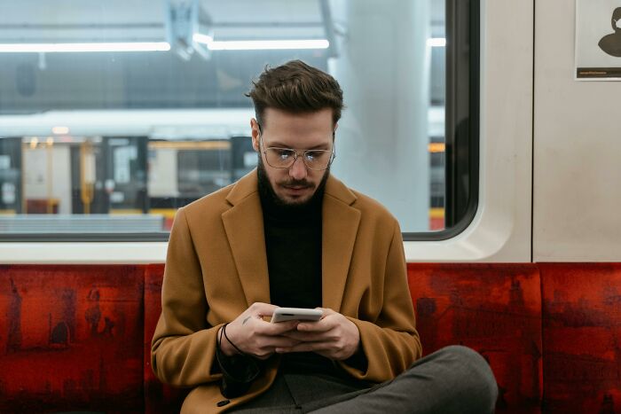 Man staying calm while using phone, seated on a train, reflecting on scenarios to see where he stands.