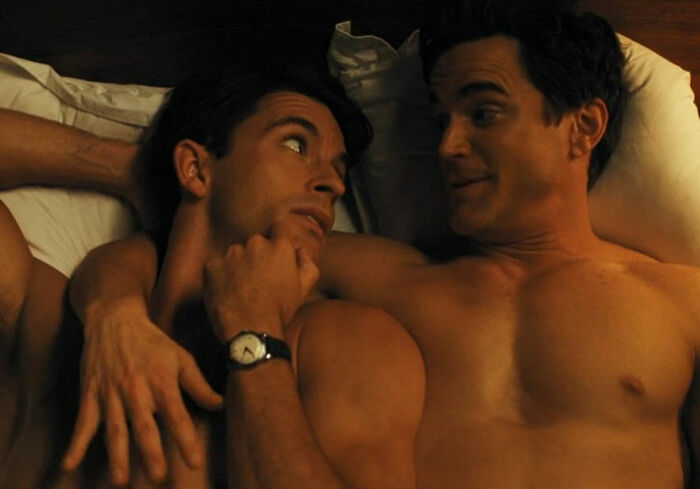 Two shirtless men lying close in bed, one looking thoughtfully at the other, evoking masculinity themes.
