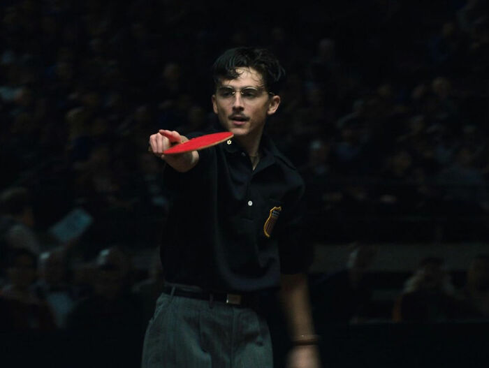 Young man in glasses playing table tennis, wearing a dark shirt and gray trousers in a dimly lit setting. Young man in glasses playing table tennis, wearing a dark shirt and gray trousers in a dimly lit setting.