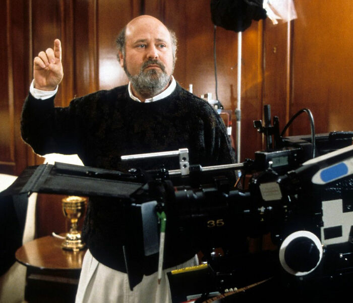 Rob Reiner directing on set, wearing a dark sweater, behind camera equipment with a wood paneled background. Rob Reiner directing on set, wearing a dark sweater, behind camera equipment with a wood paneled background.