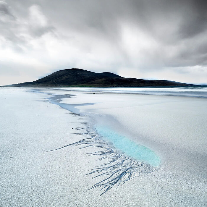 Minimalist nature photography showing a sandy beach with water patterns and a dark hill under a cloudy sky.