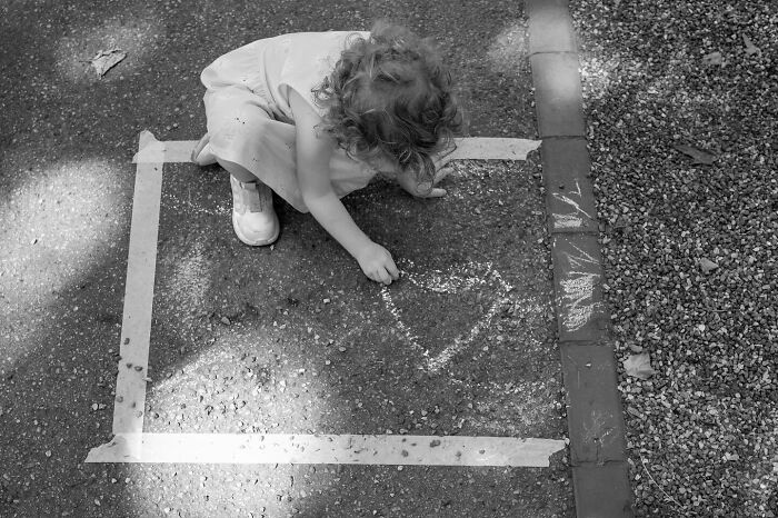 Child drawing a heart with chalk inside a taped square on the street, capturing the marvels of daily life street moments.