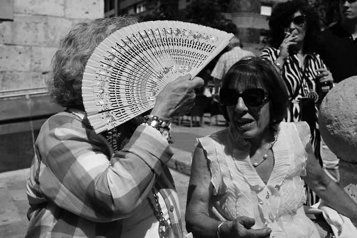 Two women in sunglasses share a moment on a city street, one shading herself with a decorative folding fan.