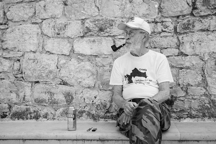 Elderly man sitting against a stone wall, smoking a pipe and enjoying a quiet street moment filled with daily life marvels.