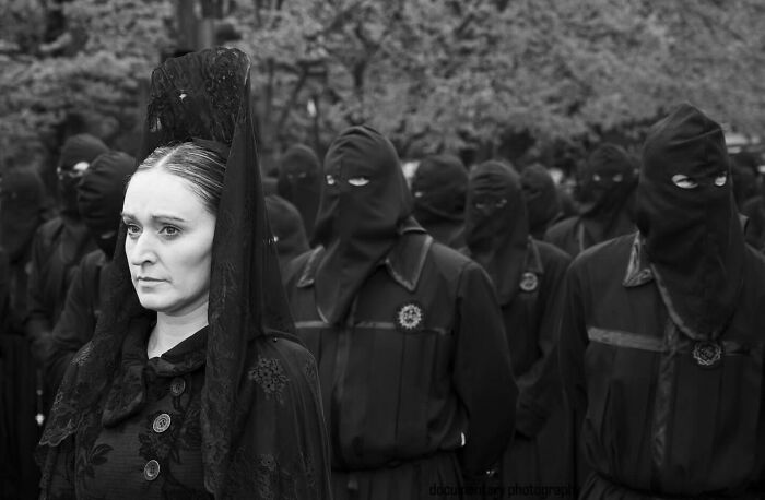 Woman in traditional black attire with lace veil standing among masked figures capturing street moments that feel like magic.