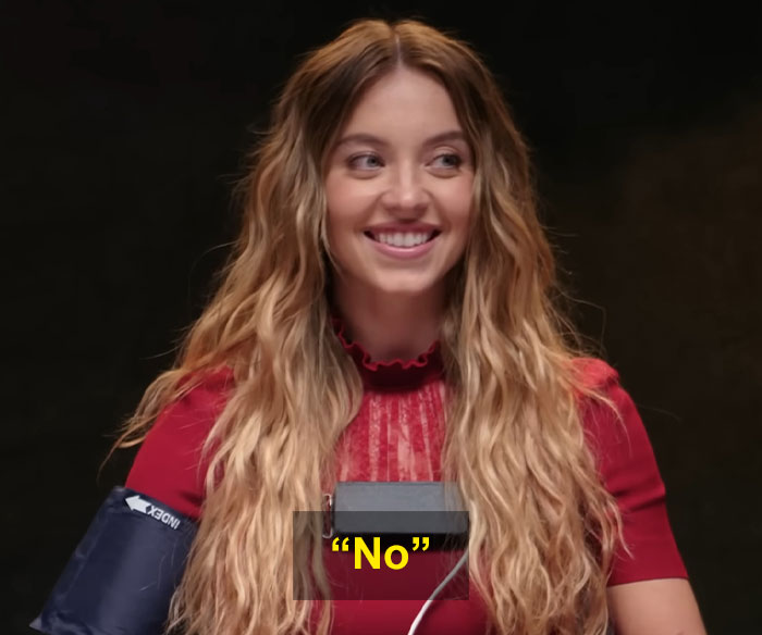 Sydney Sweeney smiling during a lie detector test while wearing a red top and medical arm cuff in a dark room. Sydney Sweeney smiling during a lie detector test while wearing a red top and medical arm cuff in a dark room.